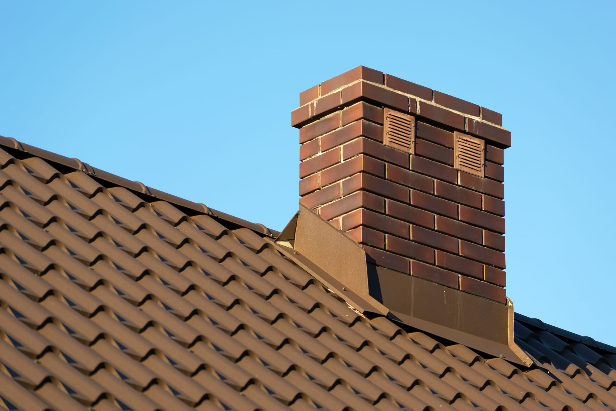 Brown brick chimney on a brown metal tile roof under a clear blue sky.