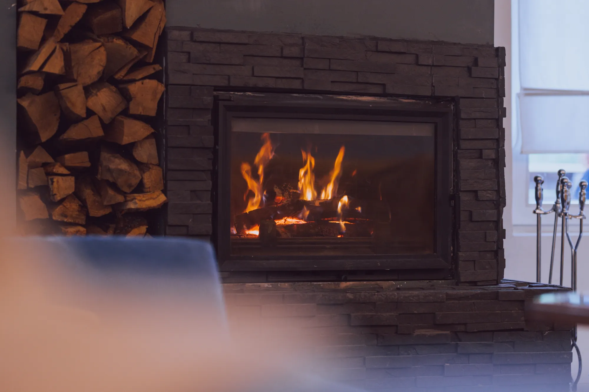A fireplace with a fire burning, next to a stack of firewood.