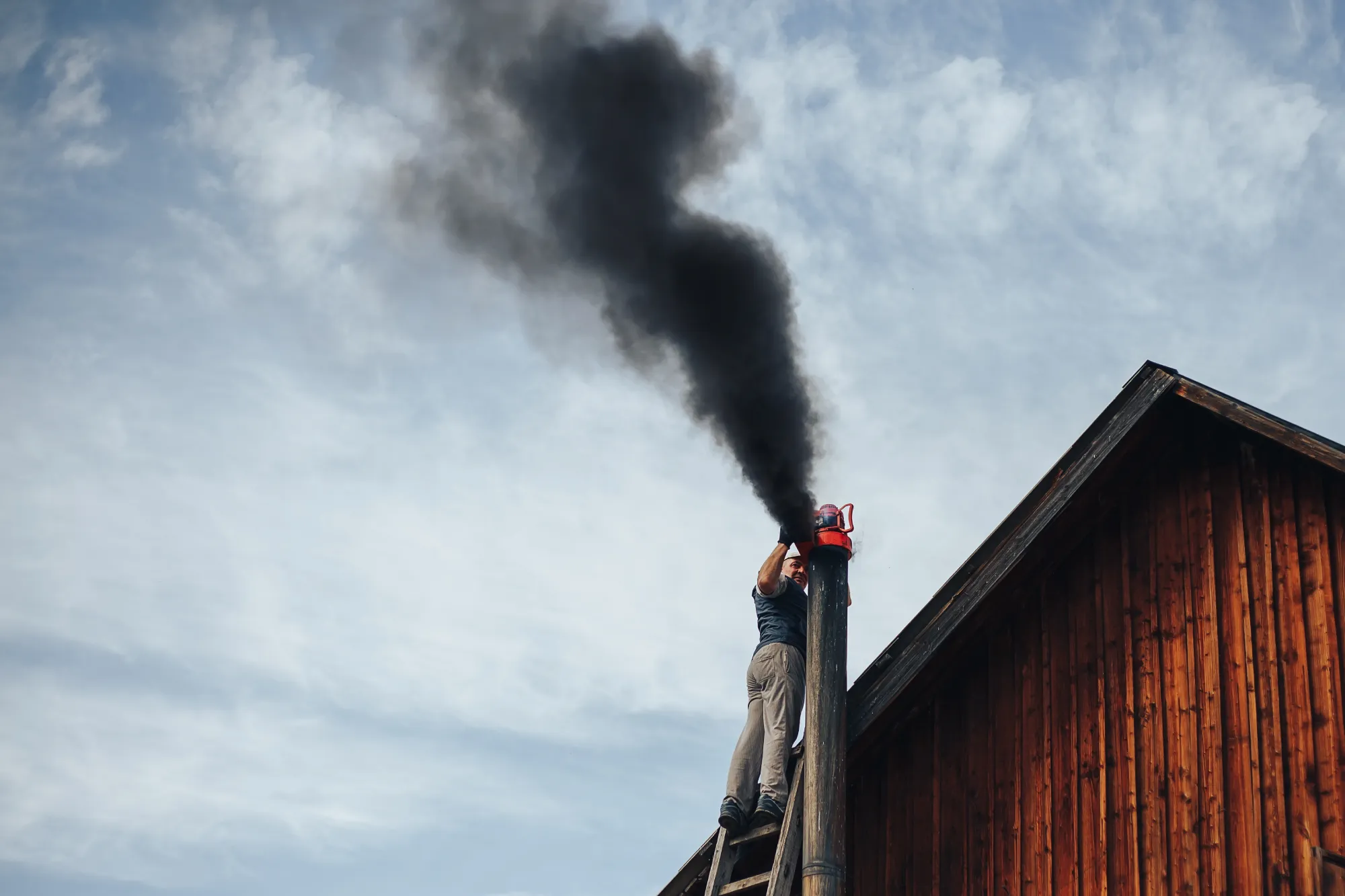 Man on ladder cleaning a smoking chimney on a wooden house.